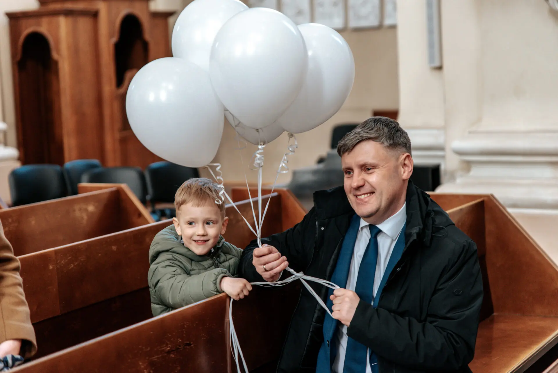 Šeimos nariai su šypsenom stebi krikštynų ceremoniją bažnyčioje, balionai pravers fotosesijoje