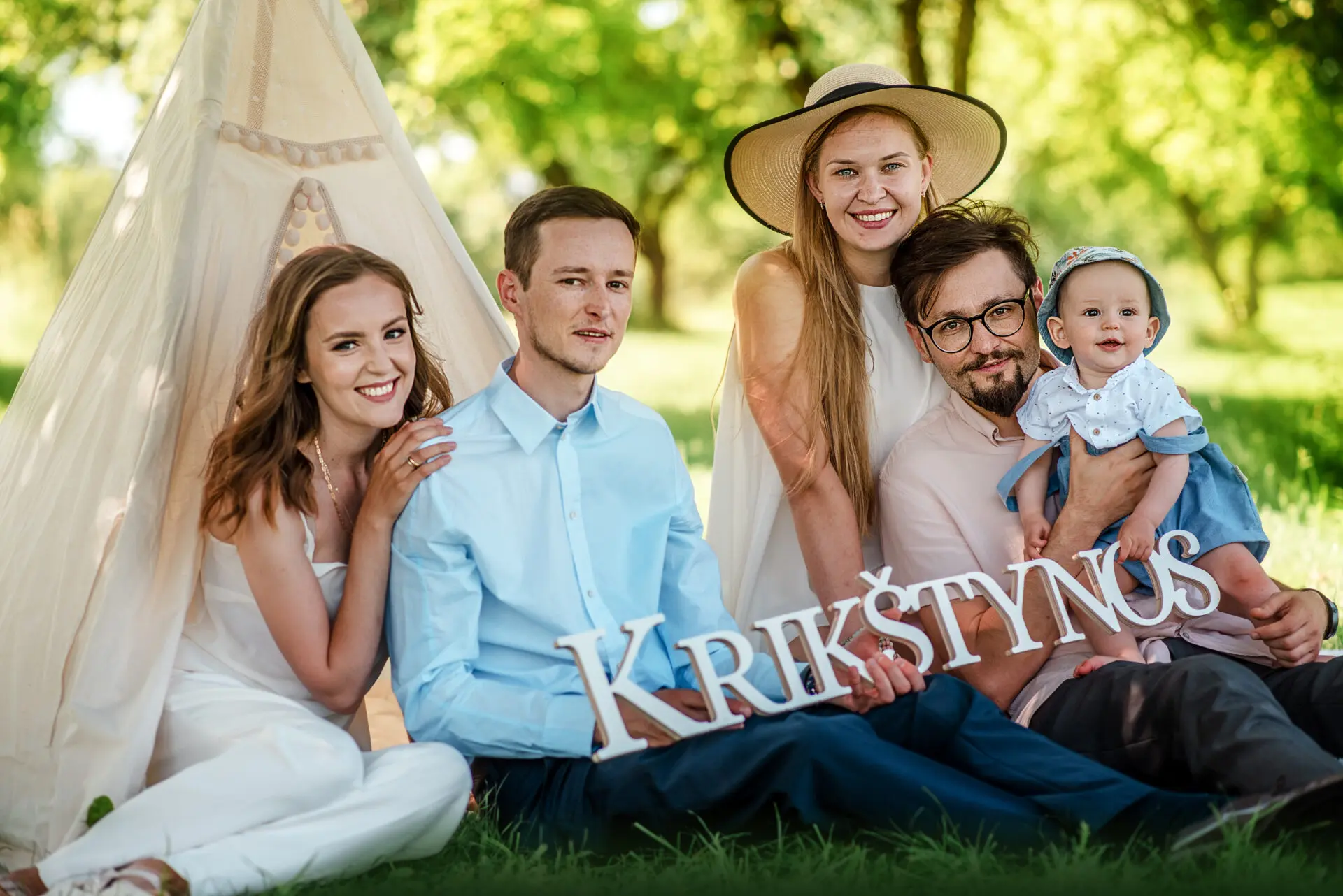 Tėvai, jų sūnus ir krikštatėviai sėdi sode ant žolės prie dekoracijų 