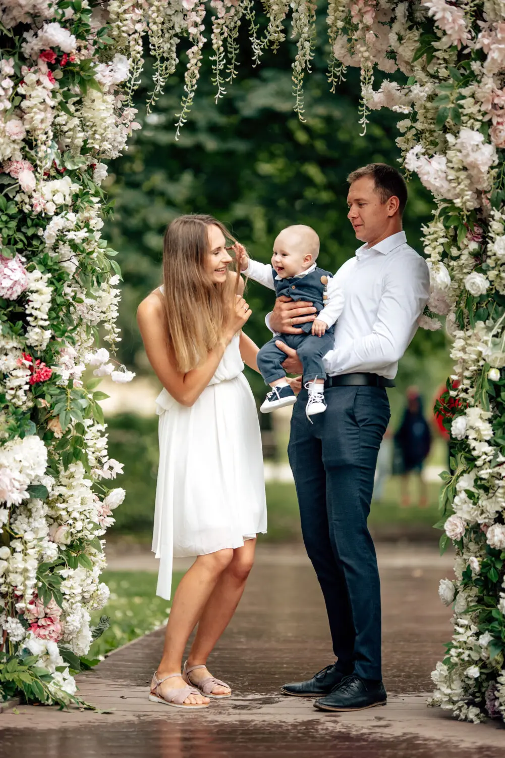 Nufotografuota šeima per krikštynų fotosesiją sode, berniukas žaismingai paima motinos plaukus, pozityvios emocijos ir gera nuotaika